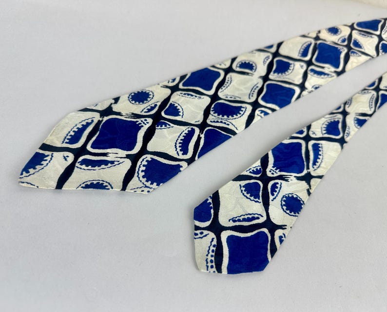 May include: A necktie featuring a repeating geometric pattern of deep blue squares with rounded corners and scalloped edges set against a cream-colored background. The design includes dark blue intersecting lines that create a grid-like structure.