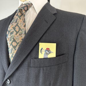 May include: A dark charcoal blazer with a white collared shirt and a patterned tie in shades of tan, green, and blue. A yellow pocket square is tucked into the chest pocket, featuring a graphic illustration of a peacock wearing a red hat.