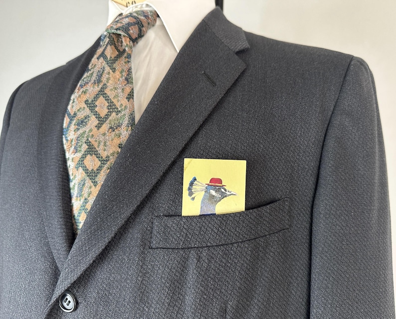 May include: A dark charcoal blazer with a white collared shirt and a patterned tie in shades of tan, green, and blue. A yellow pocket square is tucked into the chest pocket, featuring a graphic illustration of a peacock wearing a red hat.