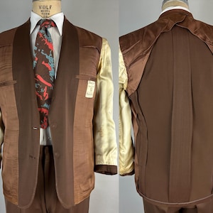 May include: A two-piece brown suit with a white shirt and patterned tie. The suit jacket has a notched lapel and two buttons. The interior lining is a light beige color. The suit is displayed on a mannequin.