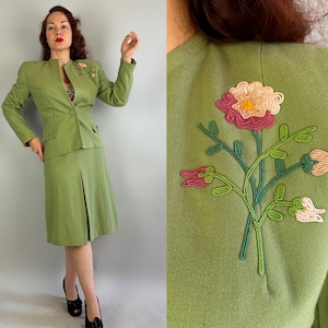 May include: A light green skirt suit with a tailored jacket and knee-length skirt. The jacket features a single-button closure, pockets, and a floral embroidered detail. The back of the jacket has a large embroidered floral design. The suit is a vintage style.