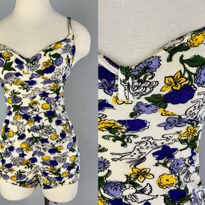 May include: A white one-piece swimsuit with a floral print in shades of yellow, blue, and green. The swimsuit has a sweetheart neckline and thin straps.