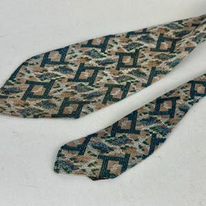 May include: A necktie featuring a repeating geometric pattern of dark teal squares on a textured background of tan, white, and muted blue. The fabric has a woven appearance with a pointed tip and a narrower tail section visible in the frame.