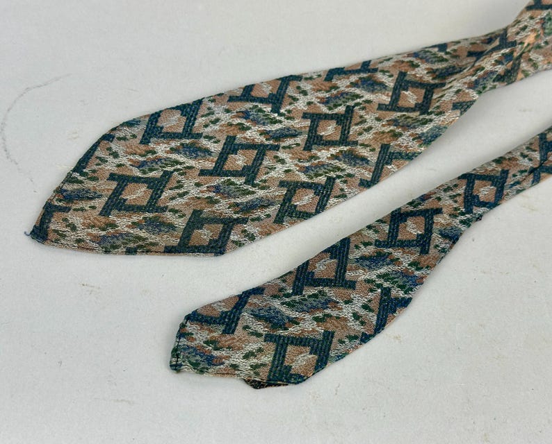 May include: A necktie featuring a repeating geometric pattern of dark teal squares on a textured background of tan, white, and muted blue. The fabric has a woven appearance with a pointed tip and a narrower tail section visible in the frame.