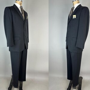 May include: A dark charcoal gray suit consisting of a two-button blazer with notched lapels and matching trousers. The blazer features a white collared shirt, a patterned tie, and a small pocket square. The suit is displayed on a mannequin.