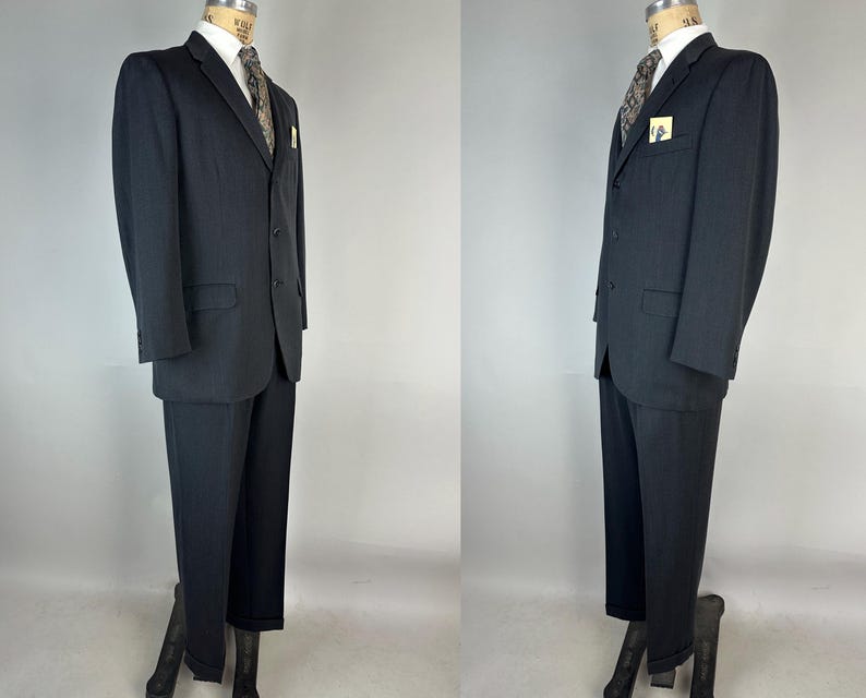 May include: A dark charcoal gray suit consisting of a two-button blazer with notched lapels and matching trousers. The blazer features a white collared shirt, a patterned tie, and a small pocket square. The suit is displayed on a mannequin.