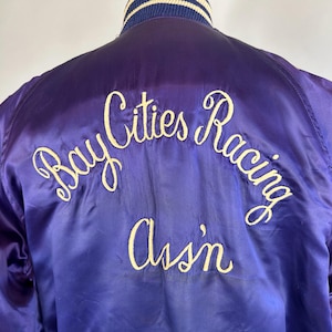 May include: A purple satin jacket with the words "Bay Cities Racing Assn" embroidered in cream-colored script across the back. The collar is striped with white and navy blue. The jacket has a classic, vintage style.
