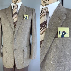 Op de afbeelding: Een lichtbruine tweed blazer met een wit overhemd en gestreepte stropdas. Een klein vierkant pochet met een pauwenillustratie zit in de borstzak. De blazer heeft twee voorzakken en een sluiting met twee knopen.