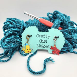 May include: A ball of teal yarn with a crochet hook and a light blue tag that says "Crafty Girl Makes". The tag features charms of a yellow fish and a red crab.