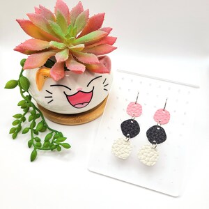 Peut inclure: Une paire de boucles d'oreilles pendantes à trois couches de simili cuir. La couche supérieure est rose, la couche intermédiaire est noire et la couche inférieure est blanche.