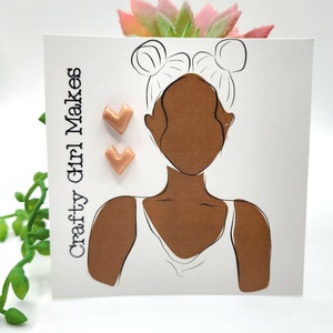 May include: A pair of rose gold stud earrings in the shape of a heart. The earrings are shown on a white card with a line drawing of a person with their hair in buns. The card also has the text "Crafty Girl Makes" on it.