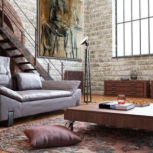 May include: A living room with a large painting of a person sitting on a chair. The painting is on a brick wall. There is a couch, a coffee table, and a rug in the room. The coffee table has a book and a glass of wine on it.