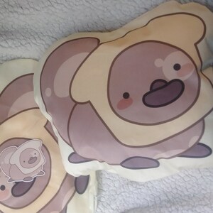 Puede incluir: Dos almohadas de felpa con forma de pingüino marrón que lleva un disfraz de pan amarillo. Las almohadas están sobre una superficie blanca y peluda.