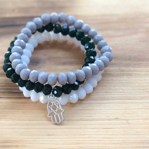 May include: Three layered bracelets with gray, white, and black beads. The bottom bracelet has a silver charm with a hamsa hand and a star of David.