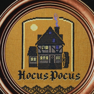 Può includere: Un ricamo a punto croce incorniciato che presenta una casa marrone scuro con una porta e finestre gialle. La casa è circondata da uno sfondo giallo con il testo "Hocus Pocus" in lettere nere.