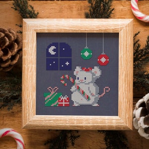 May include: Cross-stitch pattern of a white mouse holding a candy cane in front of a window with a crescent moon and stars. The mouse is wearing a red Santa hat and there are presents and ornaments around it.