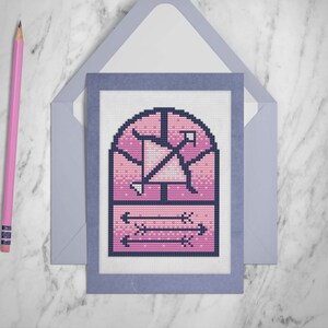 May include: A pink and blue cross-stitch card with a stained glass window design featuring arrows. The card is framed with a blue border.