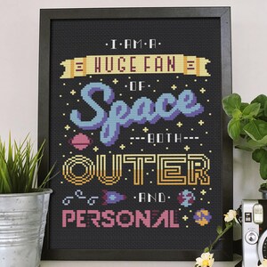 以下が含まれることがあります： 「I AM A HUGE FAN OF Space BOTH OUTER AND PERSONAL」の文字が入った、黒いフレームのクロスステッチアート。星、惑星、ロケットをカラフルなピクセルスタイルで表現した宇宙をテーマにしたデザインです。