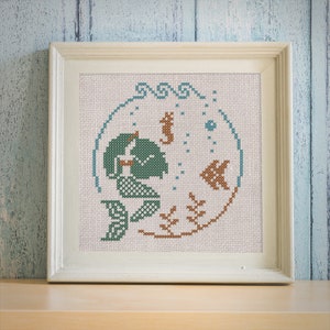 May include: A framed cross-stitch embroidery depicting a mermaid, a seahorse, and a fish in a circular design. The embroidery is done in shades of green, brown, and blue on a white background.