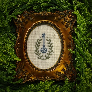 May include: A gold-colored ornate frame with a white oval mat and a cross-stitch design of a blue and green musical instrument inside.