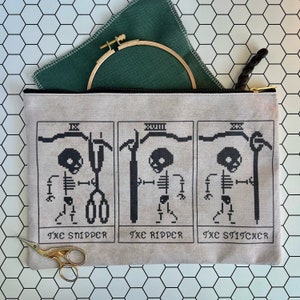 以下が含まれることがあります： 3つの異なる武器を持った骸骨の白黒刺繍が施された、ベージュのキャンバス製ジッパーポーチ。各画像の下には「The Sniper」、「The Ripper」、「The Stitcher」のテキストが書かれています。