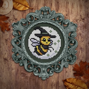 Puede incluir: Un diseño de punto de cruz enmarcado de una abeja con un sombrero de bruja. La abeja es amarilla y negra con una cara blanca y está rodeada de un fondo blanco con acentos verdes.