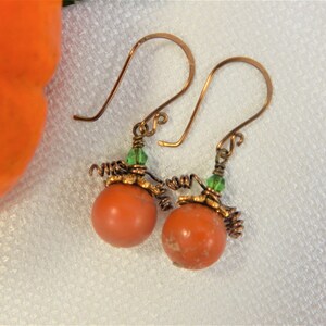 May include: A pair of orange pumpkin earrings with green crystal accents and copper wire details. The earrings are made with wire wrapped orange beads and green crystal beads.