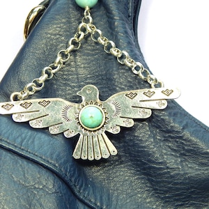 May include: A silver-toned metal bird charm with a turquoise center stone hangs from a silver chain. The charm is attached to a dark blue leather bag. The bird has detailed wing and body designs.