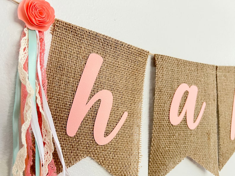 May include: A burlap banner with the word "Happy" spelled out in pink letters. The banner is decorated with pink and white ribbons and lace.