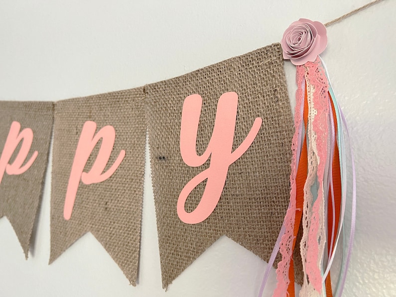 May include: A burlap banner with the word "Happy" spelled out in pink letters. The banner is decorated with a pink flower and a variety of ribbons.
