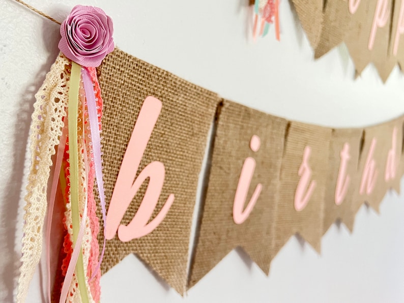 May include: A burlap banner with the word "birthday" spelled out in pink letters. The banner is decorated with a pink flower and a variety of ribbons.