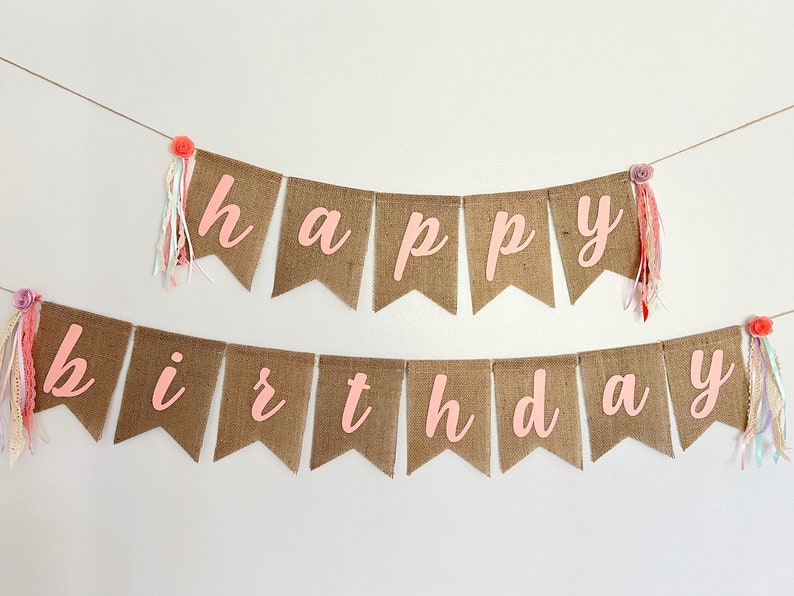 May include: A burlap banner with the words "happy birthday" in pink letters. The banner is decorated with pink and white fabric tassels and small pink flowers.