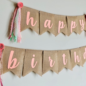 May include: A burlap banner with the words "happy birthday" in pink letters. The banner is decorated with pink and white fabric flowers and ribbons.