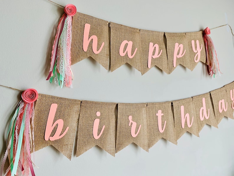 May include: A burlap banner with the words "happy birthday" in pink letters. The banner is decorated with pink and white fabric flowers and ribbons.