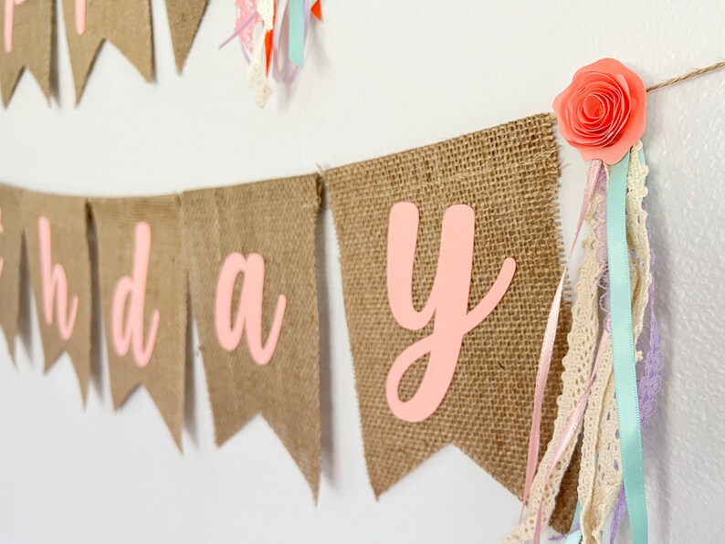 May include: A burlap banner with the word "Happy Birthday" in pink letters. The banner is decorated with pink paper flowers and ribbons.