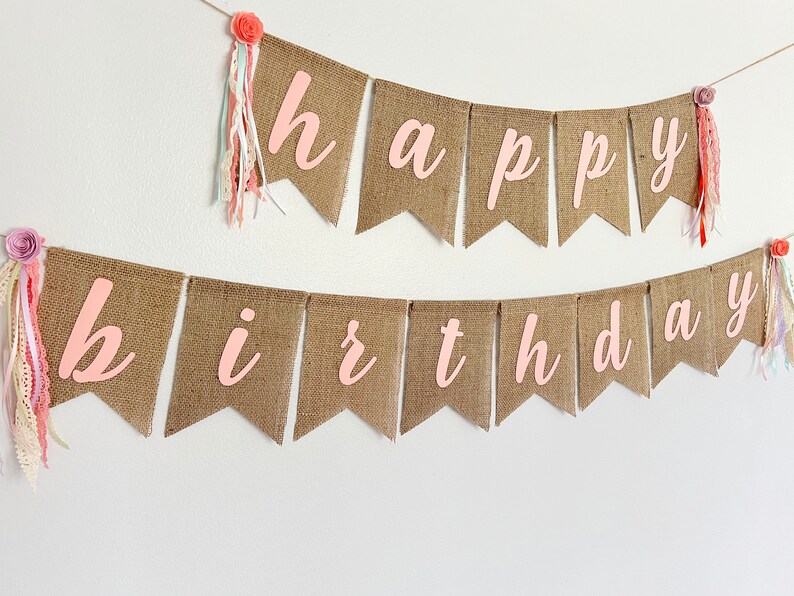 May include: A burlap banner with the words "happy birthday" in pink letters. The banner is decorated with pink and white flowers and ribbons.