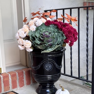 May include: A black urn overflowing with colorful fall foliage and a decorative white pumpkin with a gold stem. The arrangement includes a large green and purple cabbage-like plant, cream-colored flowers, and burgundy mums.