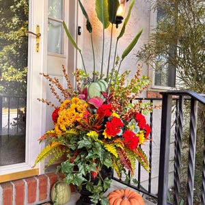 May include: A large black urn overflowing with a vibrant autumn arrangement. The display features sunflowers, red and yellow flowers, green leaves, and pumpkins in shades of green and orange. The urn is placed on a porch with a welcome mat.