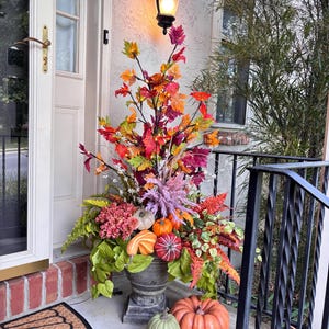 May include: A fall-themed outdoor arrangement featuring a variety of pumpkins and colorful foliage. The display includes orange, green, and striped pumpkins, complemented by vibrant red, orange, and green leaves. The arrangement is set in a decorative urn, creating a welcoming autumn scene.