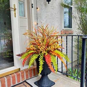 May include: A black pedestal planter filled with an arrangement of artificial autumn-themed foliage, including bright green and orange fern fronds, tall thin grasses, and small yellow sunflowers with dark centers, displayed on a porch.