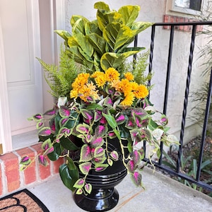 May include: A black urn-shaped planter filled with a variety of plants and flowers. The arrangement includes large green and yellow leaves, yellow flowers, and plants with pink and green leaves. The planter sits on a brick and concrete surface.