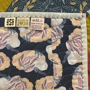 Puede incluir: Un primer plano de una colcha con fondo azul oscuro y un patrón floral. La colcha tiene un borde amarillo y una etiqueta que dice "MADE IN 2024" y "LOOK AFTER ME".