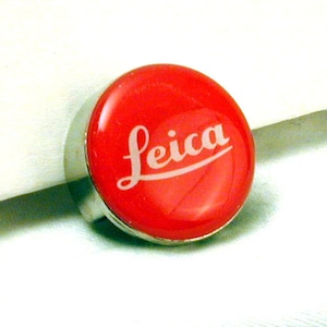 Pode incluir: Um botão redondo vermelho com uma borda prateada e a palavra "Leica" em escrita branca.
