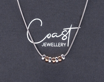 May include: A silver chain necklace with a row of small silver and gold beads. The necklace is displayed in a gray box with the text "Coast Jewellery".