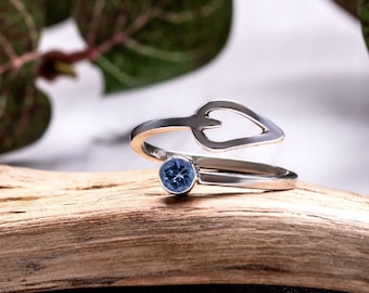 May include: A silver ring with a blue gemstone and a leaf design. The ring is on a piece of driftwood.