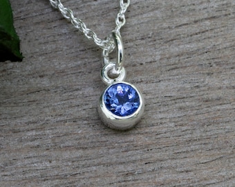 May include: A delicate silver necklace featuring a small, round, bezel-set blue gemstone pendant. The pendant is suspended from a fine silver chain. The background is a wooden surface with a green leaf.