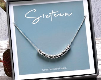 May include: A silver necklace with a row of small beads is displayed in a gift box. The box has a blue background with the word "Sixteen" in cursive. The box is on a wooden surface.