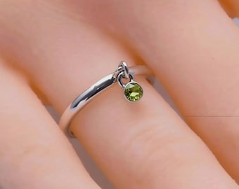 May include: A silver ring with a small, round, light green gemstone dangling from a loop. The ring is simple and elegant, with a polished finish. The gemstone is a vibrant green color.