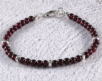 May include: A beaded bracelet featuring small, round, deep red beads. The bracelet has silver-colored spacers and a silver clasp. The beads are strung together to form a complete circle. The bracelet is displayed on a textured, white surface.