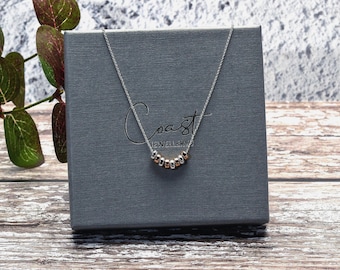 May include: A silver necklace with a series of small, alternating silver and rose gold beads. The necklace is displayed on a gray box with the word "Coast" printed on it. The box is sitting on a wooden surface.